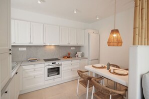 A modern kitchen with white cabinetry, a built-in oven, a dining area with a table and wicker chairs, and a hanging light fixture. - Malvia Loft Kastraki Naxos, Brand New within walking distance to the beach. (Kastraki)