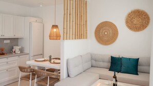 A modern kitchen with white cabinetry, a dining area with a white table and wicker chairs, and a living area with a grey sofa and teal pillows.