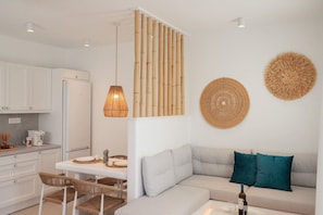 A modern kitchen with white cabinetry, a dining area with a white table and wicker chairs, and a living area with a grey sofa and teal pillows.