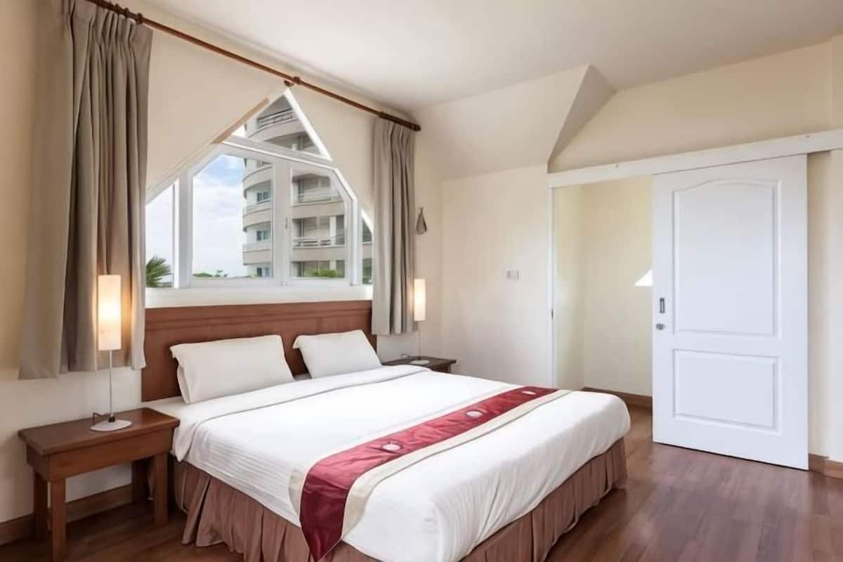 A hotel room with a large bed, a nightstand, a window with curtains, a door, and a view of a building outside.