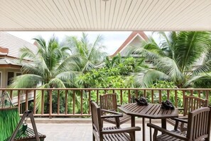 A covered outdoor seating area with wooden furniture, a view of a tropical landscape with palm trees, and a building with a red roof in the background. - Villa21Chaam (Cha-am)