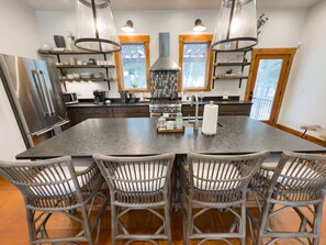 Una cocina con una isla grande, electrodomésticos de acero inoxidable, sillas de mimbre e iluminación colgante.