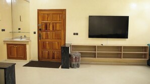 A modern living room with a wooden door, a large flat-screen TV mounted on the wall, a marble accent piece, and a wooden entertainment unit. - Almouahidine Residence (Fes)