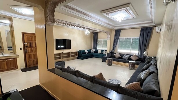 A spacious living room with a large sectional sofa, a flat-screen TV, and a kitchen area visible in the background. - Almouahidine Residence (Fes)