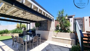 Ein Außenbereich mit Terrasse, ausgestattet mit einem Tisch und Stühlen mit Glasplatte, einem an der Wand befestigten Fernseher und einer Treppe zu einer überdachten Terrasse.