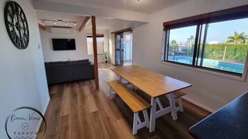 A modern living room with a wooden dining table, a grey sofa, a flat-screen TV, and a large window overlooking a pool.