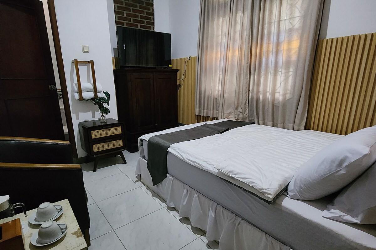 Habitación de hotel con cama, televisor, armario, una mesita con tazas y ventana con cortinas.
