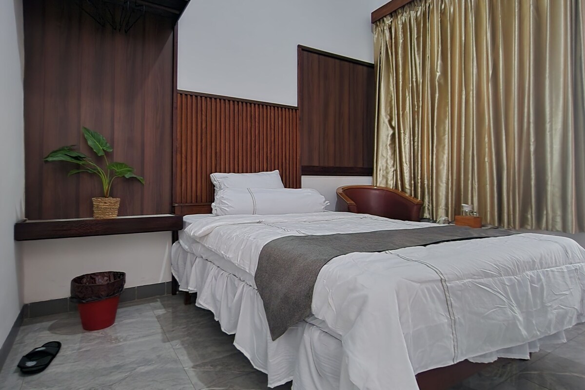 Een slaapkamer met een bed, een raam met gordijnen, een plant en een prullenbak.