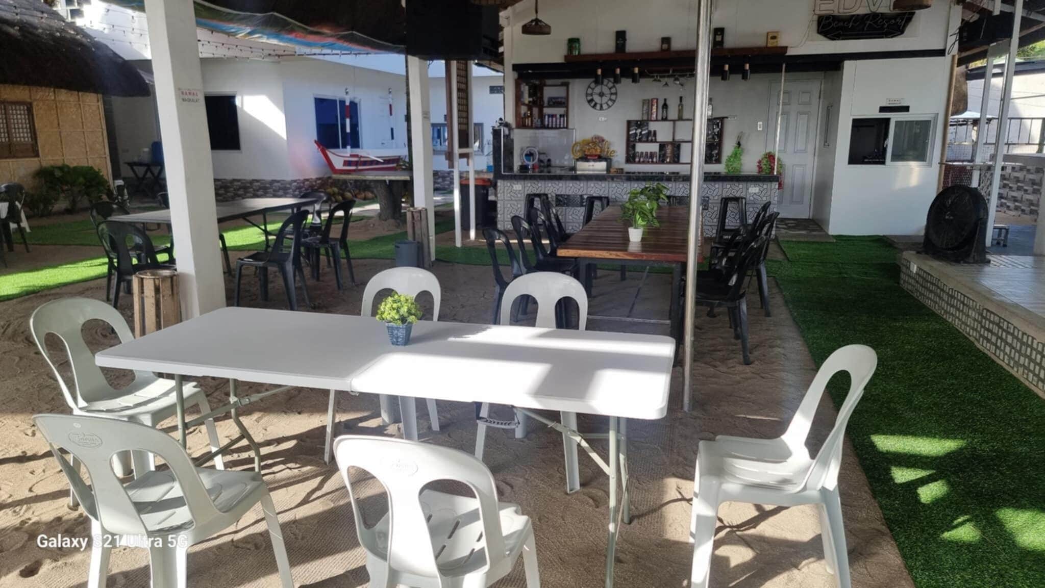 An outdoor dining area with a white table and chairs, surrounded by a covered patio with a bar area in the background.