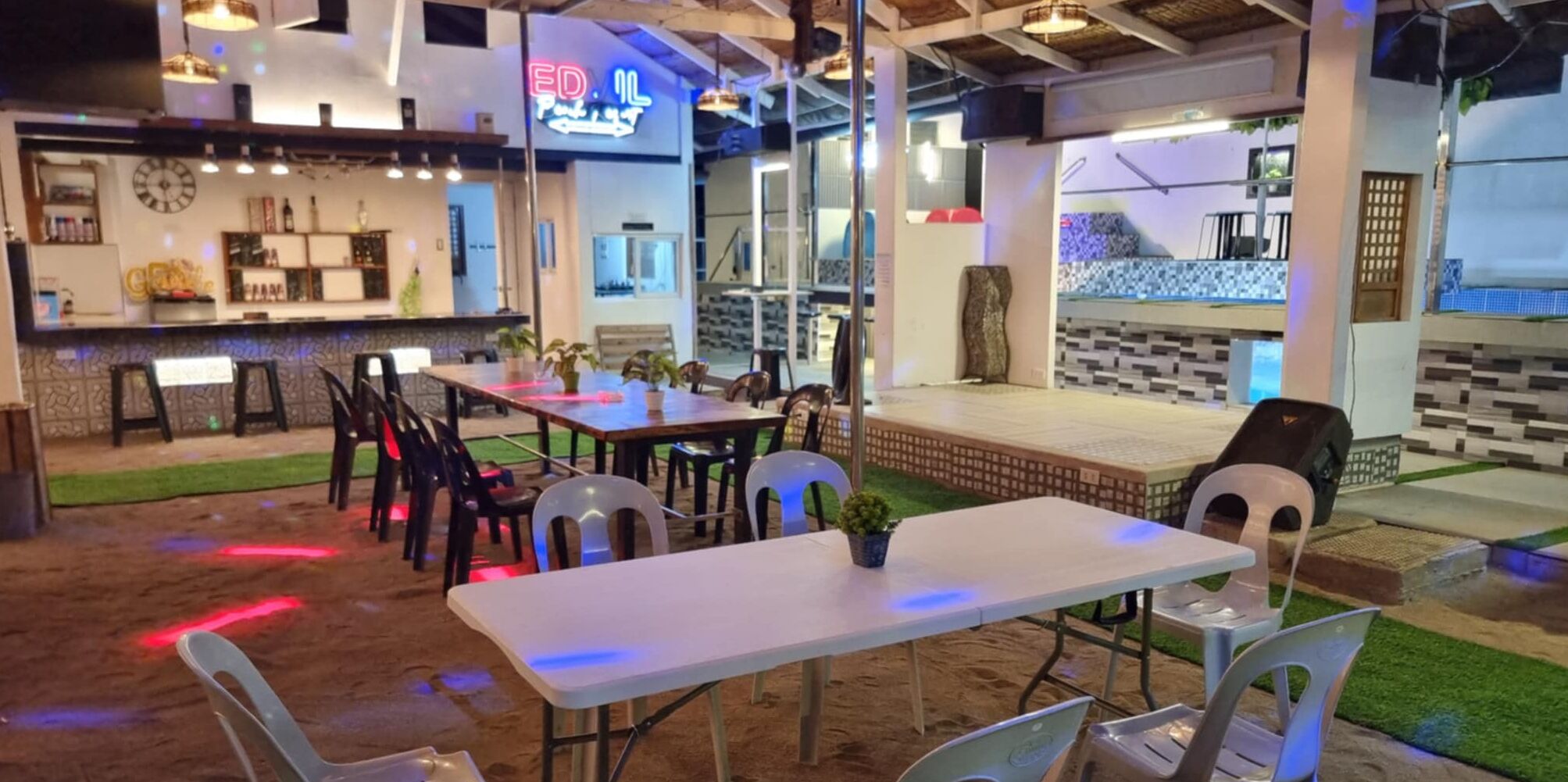 An indoor dining area with tables and chairs, a bar area with a neon sign, and a view of the outside.