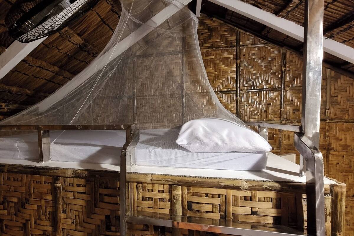 Una habitación rústica con techo de paja, una cama con almohada blanca y un muro tejido.
