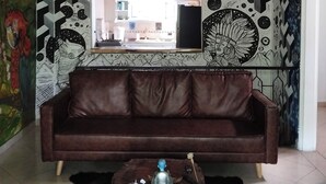 Une pièce avec un canapé en cuir marron, une table basse en bois, une fresque murale aux motifs géométriques et une vue sur une cuisine par une fenêtre.
