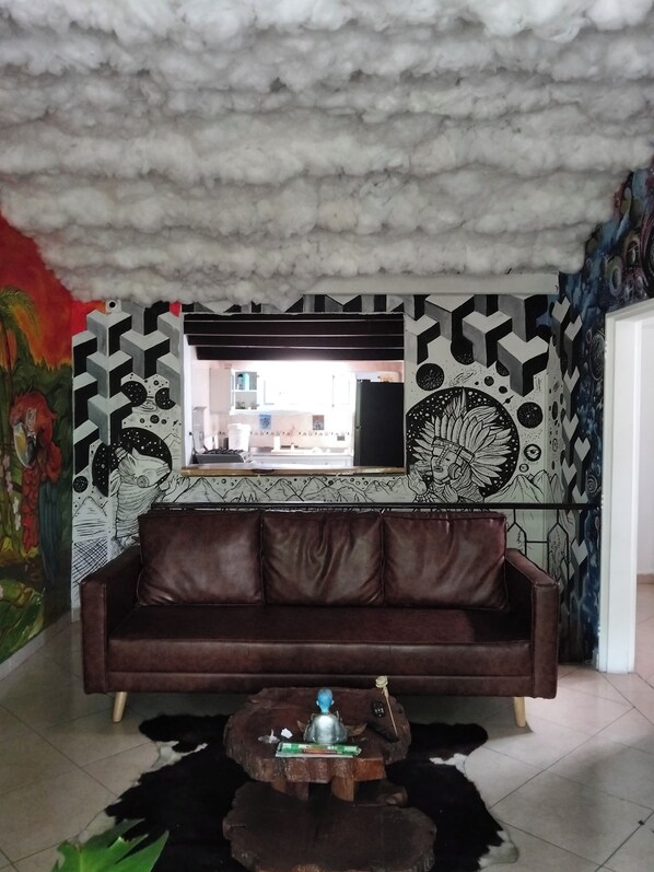 A room with a brown leather sofa, a wooden coffee table, a mural with geometric patterns, and a view of a kitchen through a window. - Nature Oasis Near Provenza, Private room for two. In front of beautiful park!  (Medellín)
