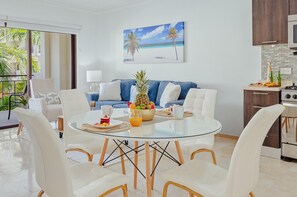 Uma sala de estar moderna com uma mesa de jantar de vidro, sofás brancos e uma área de cozinha com fogão e armários.