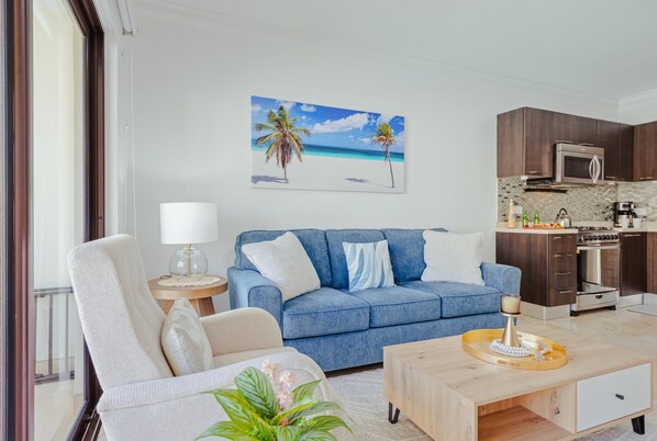 A living room with a blue sofa, a wooden coffee table, a white armchair, and a lamp. There is a kitchen area with a microwave, stove, and cabinets. A painting of a beach scene hangs on the wall.