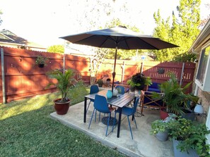 Een terras met een houten tafel en blauwe stoelen, omgeven door potplanten en een houten hek.