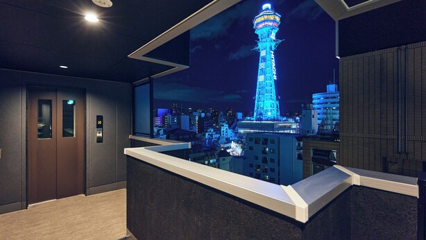 A modern hotel lobby with an elevator, a reception desk, and a cityscape view featuring a lit tower. - MONTANA WING YEBISU (Osaka)
