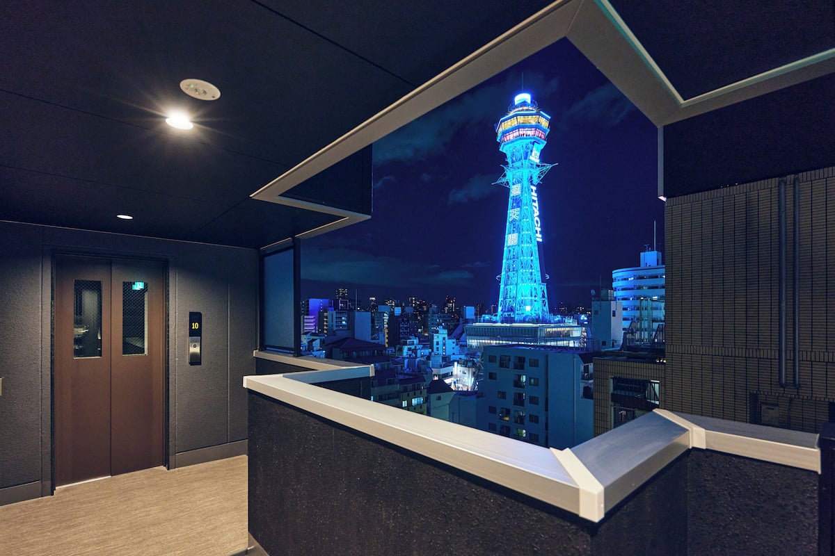A modern hotel lobby with an elevator, a reception desk, and a cityscape view featuring a lit tower.