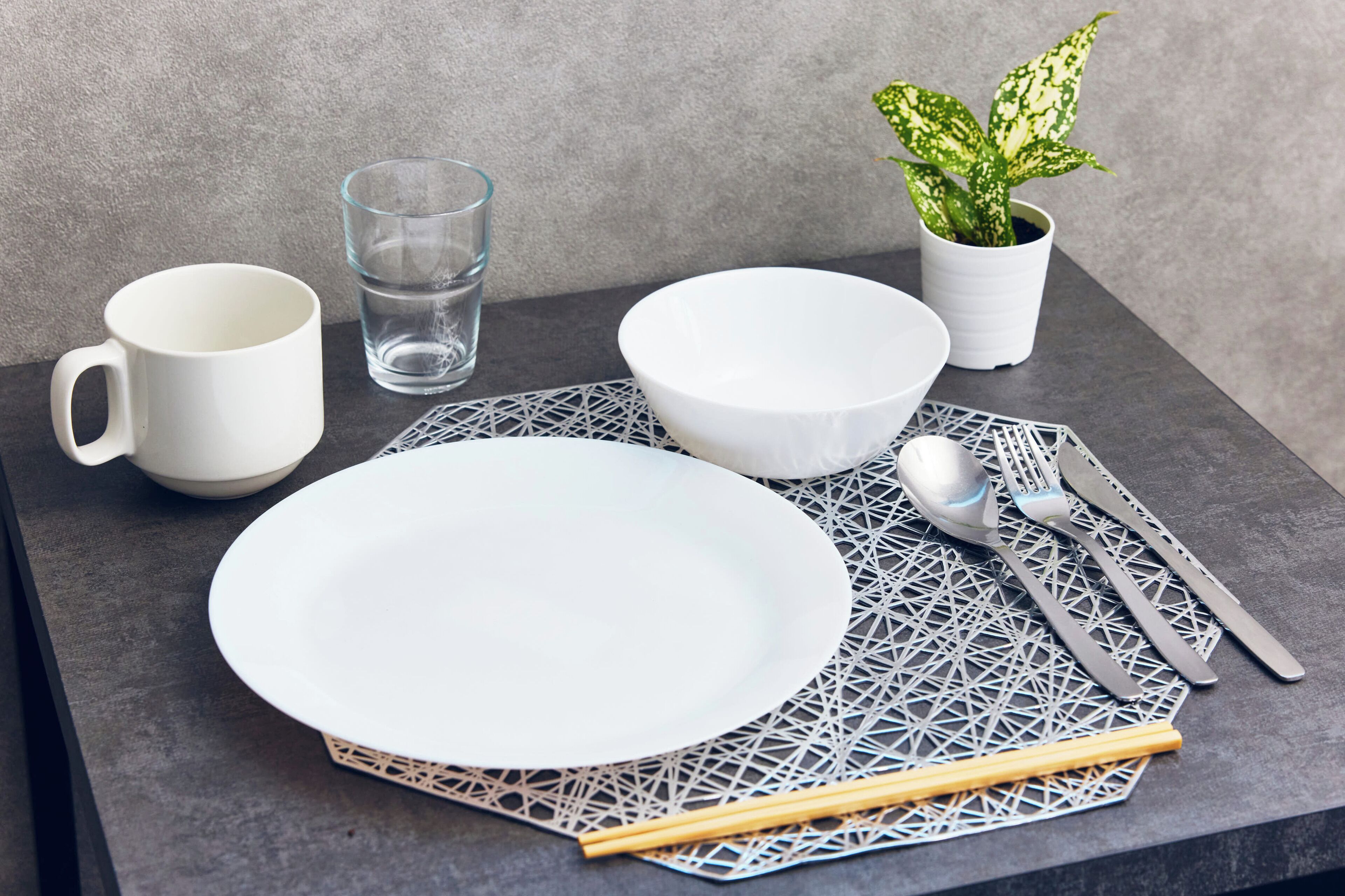 Una mesa dispuesta con vajilla blanca, un vaso de agua, una planta en maceta y palillos.