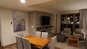 Ein modernes Wohnzimmer mit einem Holzesstisch, grau gepolsterten Stühlen, einem flachbildfernseher an der Wand und einer Sofagarnitur.