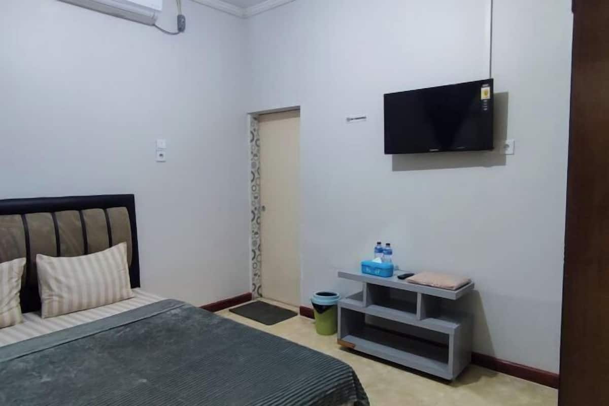 A bedroom with a bed, a TV mounted on the wall, a small table with a water bottle, and a trash bin.