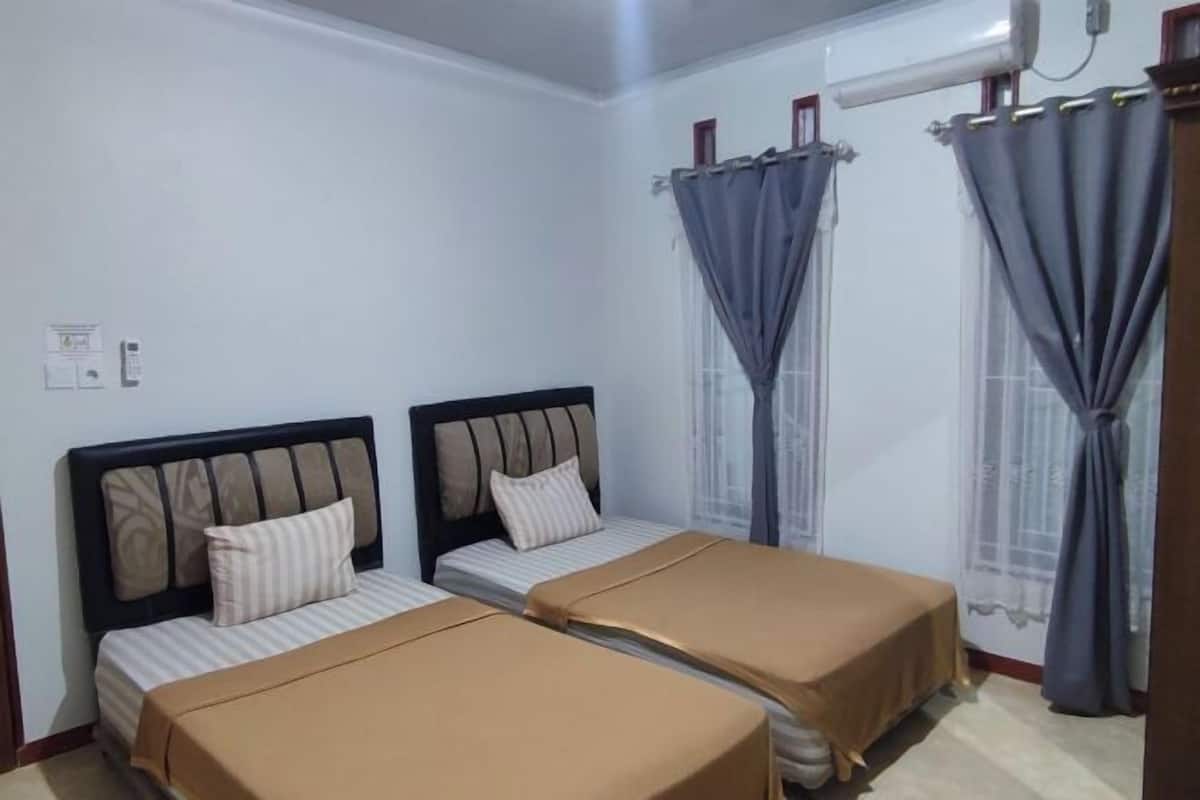 A room with two beds, a window with curtains, a wall-mounted air conditioner, and a light switch.
