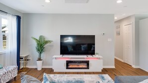Una sala moderna con una televisión de pantalla plana sobre un mueble blanco, una chimenea y un gran ventanal con cortinas.