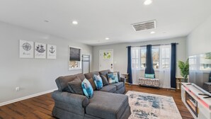 Una sala de estar con un sofá seccional gris, una mesa de centro y un mueble para televisión. Hay tres cuadros en la pared y una ventana con cortinas.