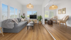 Un salón amplio con un sofá gris, una mesa de centro, sillas de mimbre y un televisor sobre un mueble de madera.