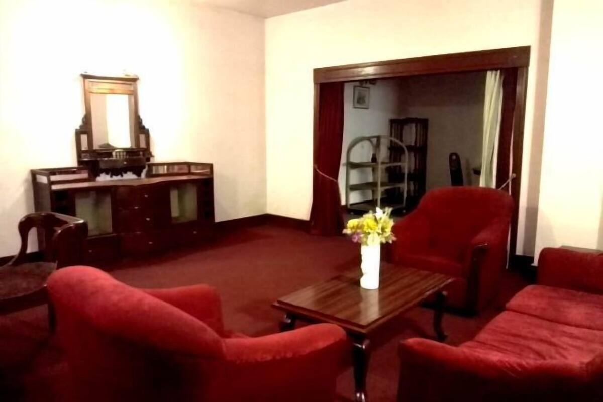 Quarto com escrivaninha de madeira, espelho, poltrona vermelha, mesa de centro de madeira com um vaso de flores e sofá vermelho.