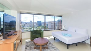 Una habitación de hotel moderna con un ventanal grande, una cama, un escritorio con televisor y un sillón verde.