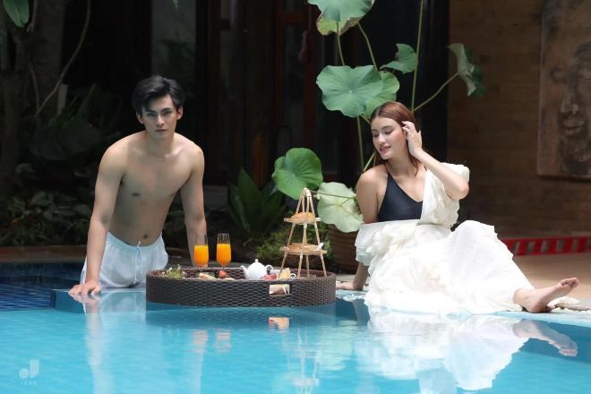 Un homme et une femme sont assis au bord d’une piscine, devant une table où est préparé un petit-déjeuner.