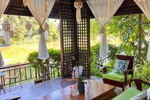 A covered outdoor seating area with a table and chairs, a decorative plant, and a view of a natural landscape. - Luxurious Living by the River Hannah Villa (Chiang Mai)