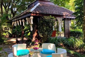 A gazebo with a tiled roof, surrounded by lush greenery and wicker furniture. - Luxurious Living by the River Hannah Villa (Chiang Mai)