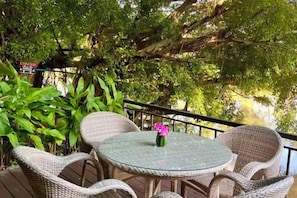 An outdoor patio with wicker furniture and a table, surrounded by greenery and a railing. - Luxurious Living by the River Hannah Villa (Chiang Mai)