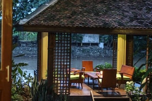 Un espace extérieur aménagé avec des meubles en bois, comprenant une table et des chaises, entouré de plantes et offrant une vue sur une rivière.