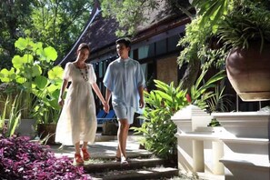 Two people walking on a pathway surrounded by plants and a building with a traditional roof. - Luxurious Living by the River Hannah Villa (Chiang Mai)