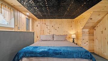Una camera da letto con soffitto in legno, un letto con coperta blu e un comodino con lampada.
