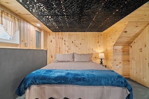 Un dormitorio con techo de madera, una cama con manta azul y una mesita de noche con lámpara.