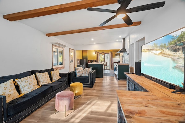 Una sala de estar moderna con un sofá negro, un reposapiés rosa y una mesa de centro de madera. Una pantalla grande de televisión muestra una vista panorámica del exterior.