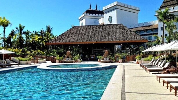 Un área de piscina en un resort, con una piscina grande, sillas de descanso y un edificio de techo de paja.