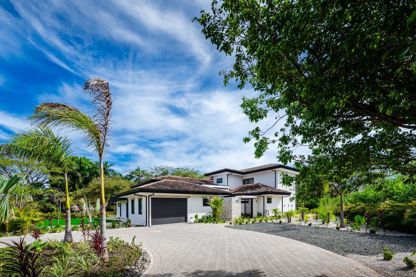 A modern house with a paved driveway, surrounded by lush greenery and a clear blue sky.