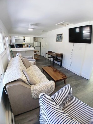 A compact living space with a sofa, a striped chair, a wooden coffee table, a dining table with chairs, a kitchen area, and a TV mounted on the wall.