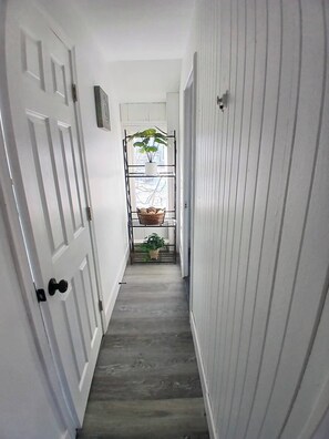 A narrow hallway with white walls and a wooden floor, leading to a window with a plant and a basket.