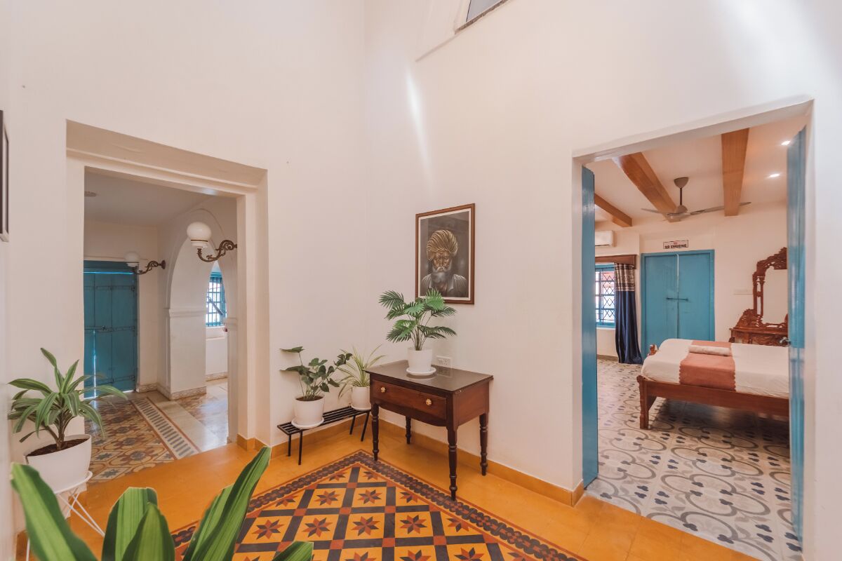Un couloir avec un tapis à motifs, un bureau en bois orné d’une plante en pot, un tableau encadré accroché au mur, et un lit avec une tête de lit bleue dans la pièce adjacente.