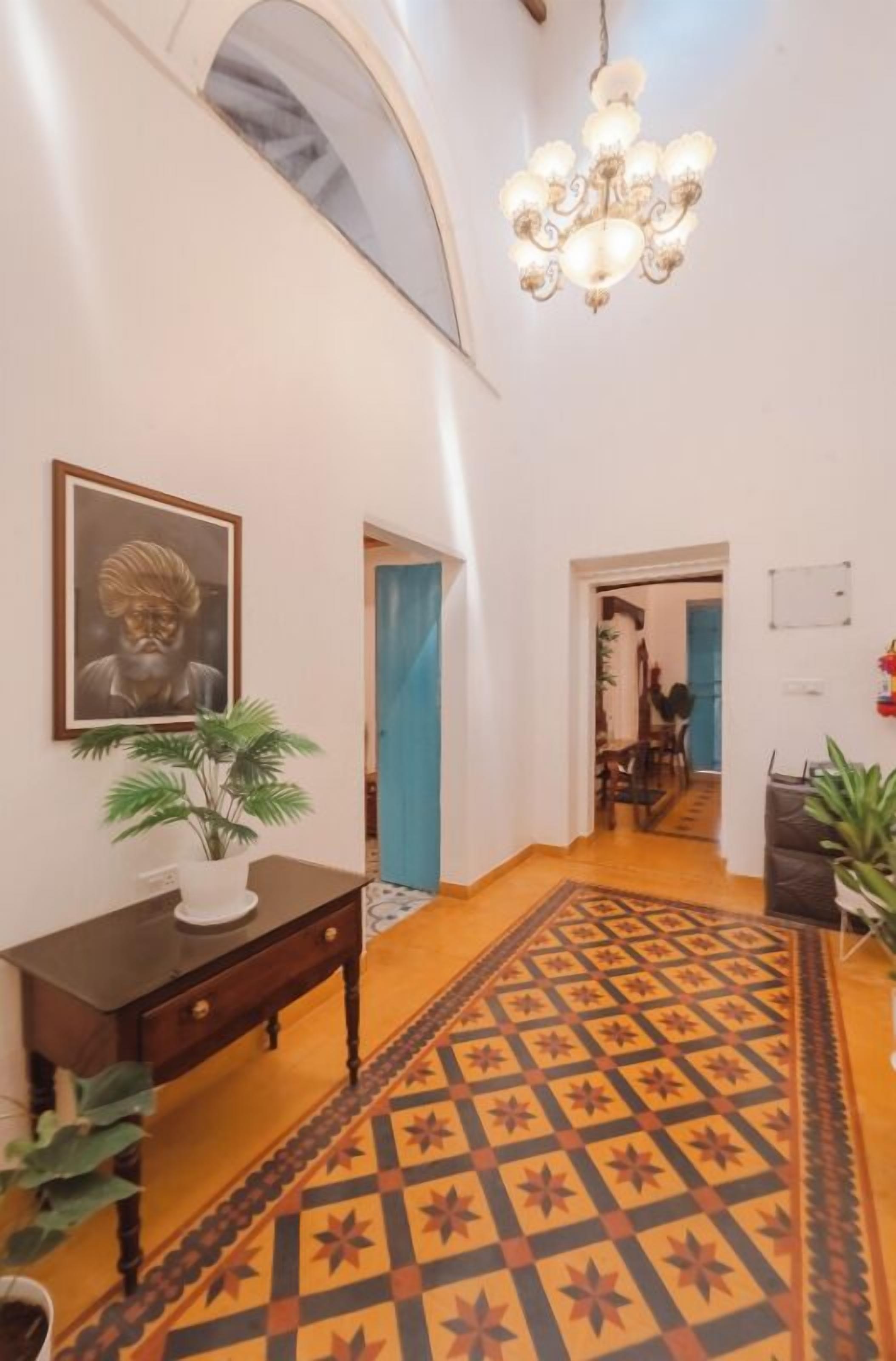 A hallway with a patterned carpet, a chandelier, a framed picture, and a potted plant.