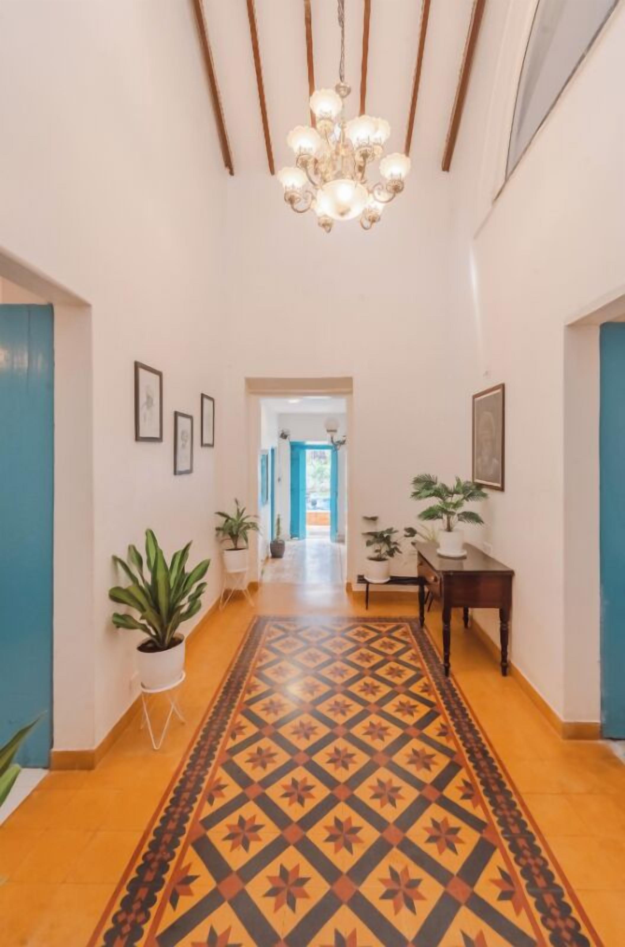 A hallway with a patterned carpet, a chandelier, potted plants, framed pictures on the walls, and a wooden table.