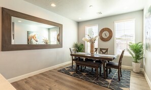 A dining area with a wooden table, chairs, a large mirror, a wall clock, and potted plants.