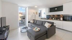 Een modern appartement met een grijze bank, een eettafel met een dienblad met kopjes en een kaars, en een keuken met witte kasten en een zwart aanrecht.