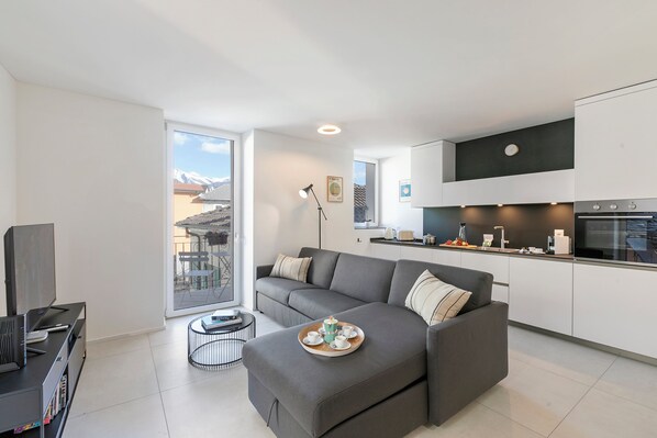 Un apartamento moderno con un sofá gris, una mesa de comedor con una bandeja de tazas y una vela, y una cocina con armarios blancos y una encimera negra.